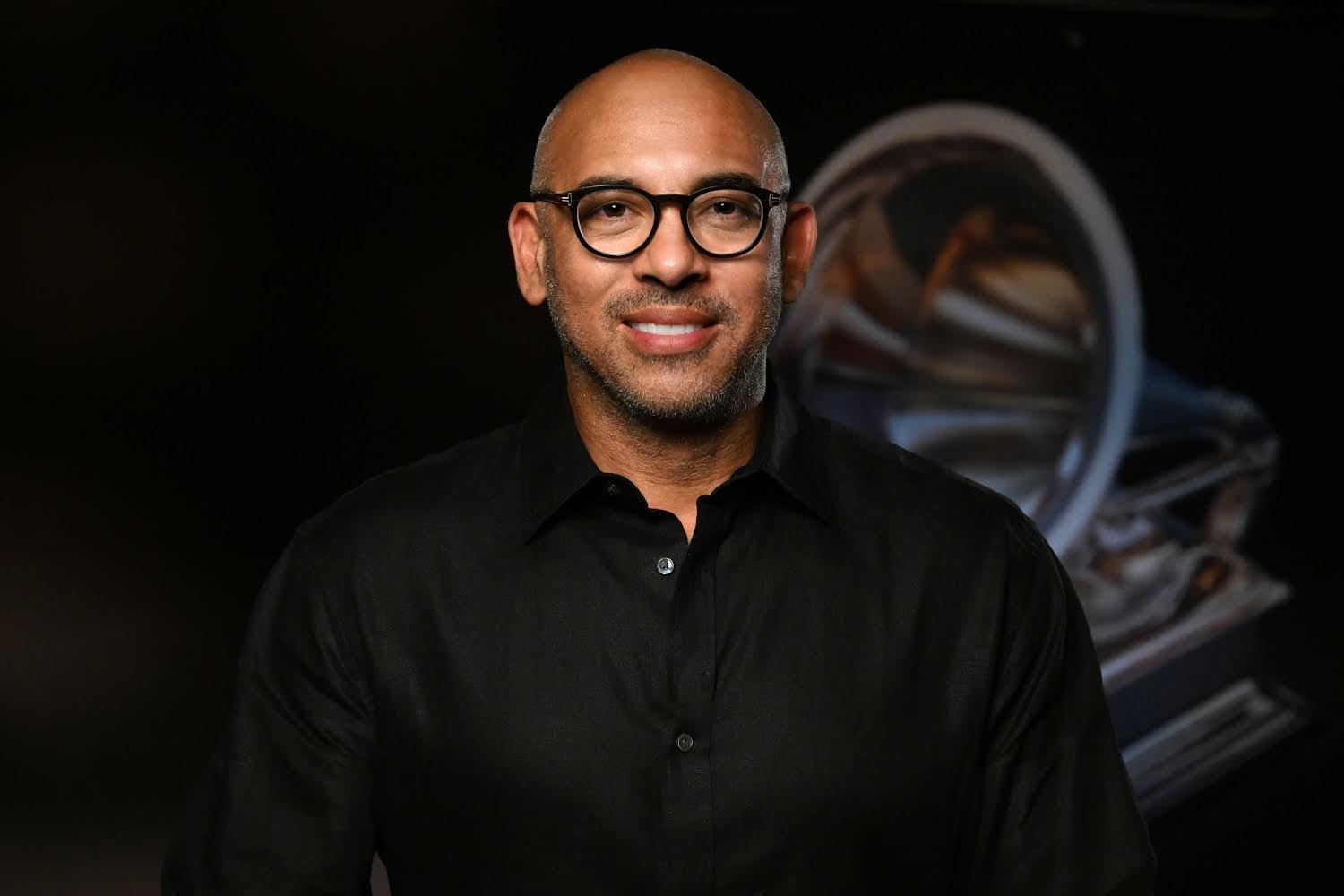 Photo of Recording Academy CEO Harvey Mason jr. smiling and wearing a black long-sleeve shirt and black-rimmed glasses against a background featuring a GRAMMY Award